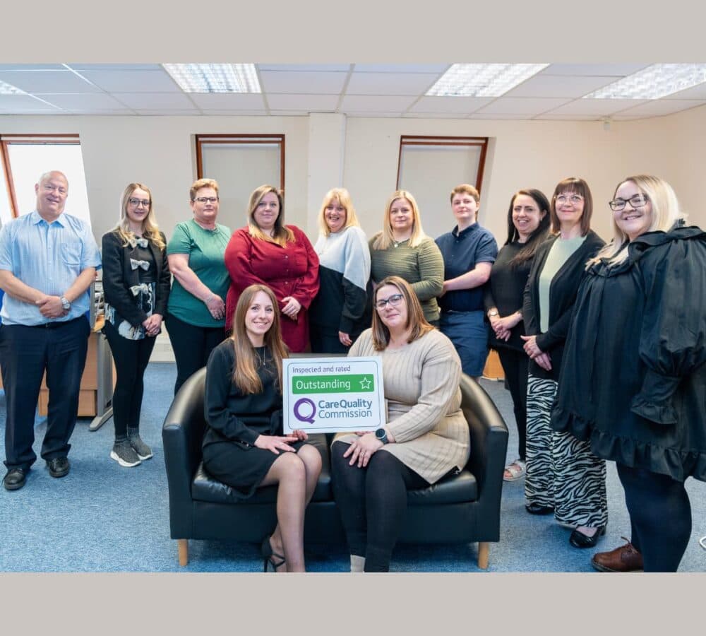 Group of people all happy and smiling inside the office with two people sitting and holding Outstanding Care Quality Commission Award