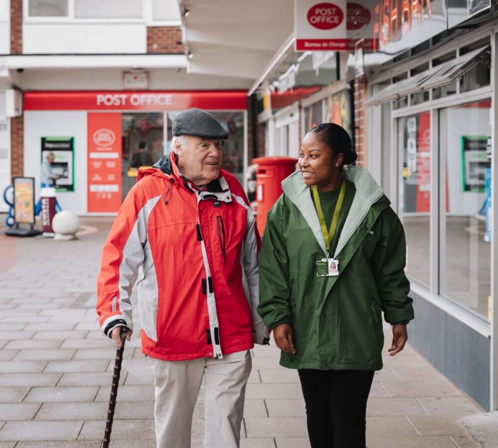 An older male adult wearing a hat and sweater and using a crutch while walking with his younger female carer with black hair and wearing a sweater both chatting while walking