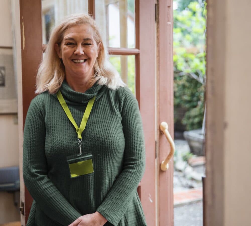 Smiling woman with a name badge stands in a doorway, wearing a green sweater, with greenery outside. - Home Instead