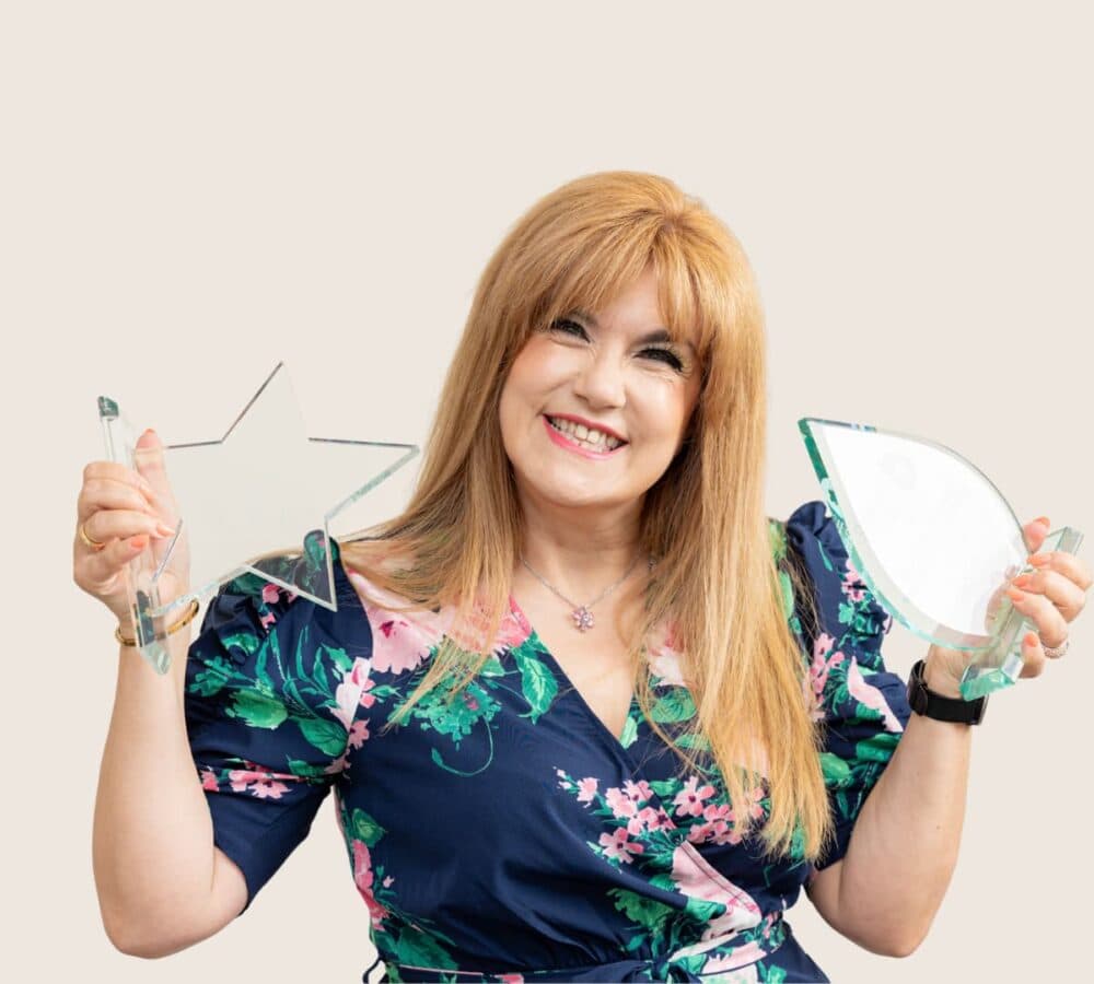 Woman with long blonde hair and wearing green floral dress smiling while holding two awards