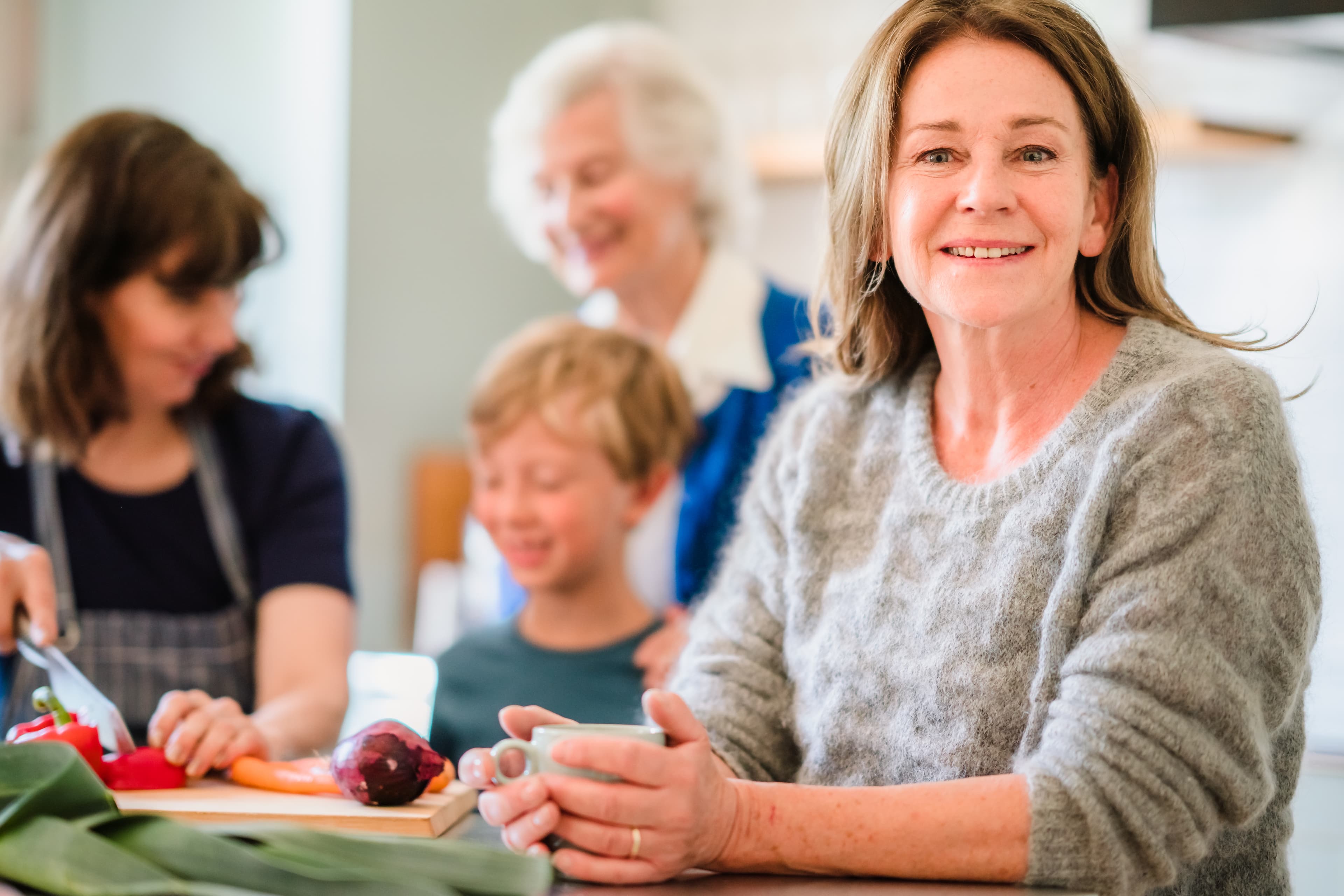 Woman smiling at the camera while family members prepare food together in a bright kitchen. - Home Instead