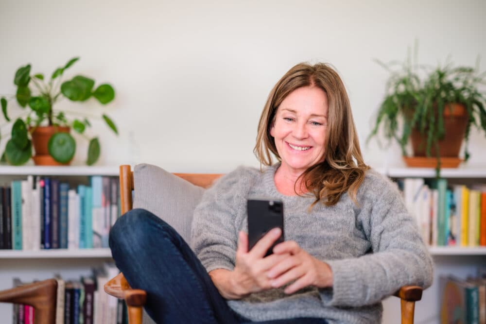 Woman sitting on a chair, smiling at her phone, with bookshelves and plants in the background. - Home Instead