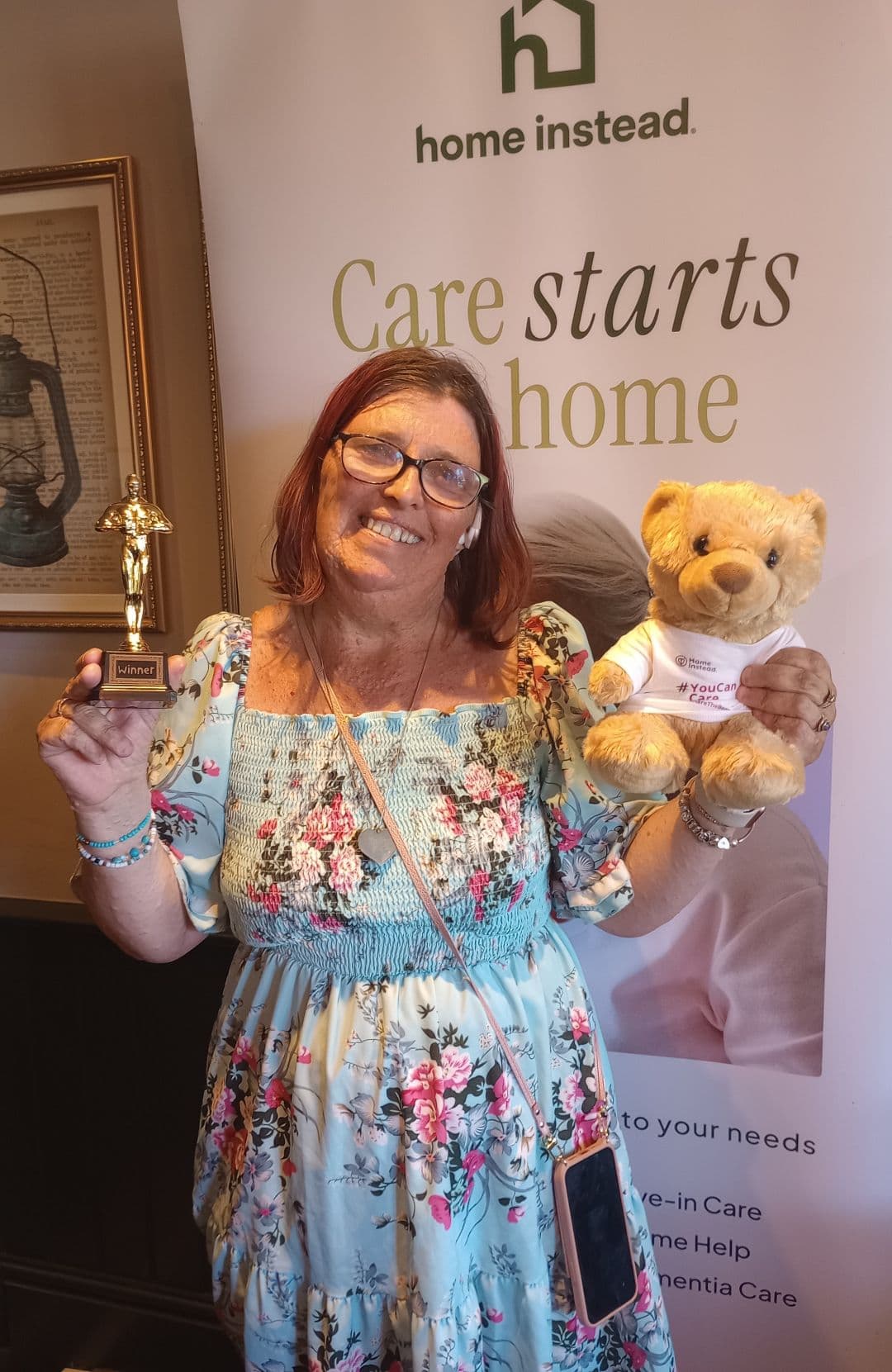 Smiling woman in a floral dress holds a trophy and teddy bear in front of a "Care starts at home" banner. - Home Instead