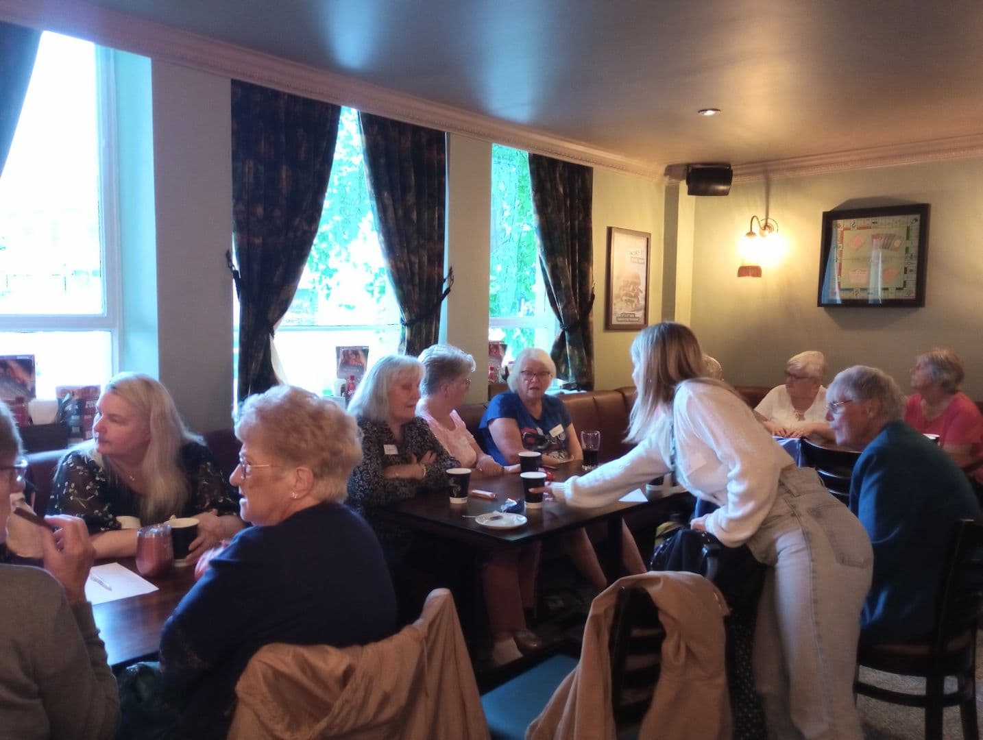 A group of older women sit chatting at tables in a cozy, well-lit pub or café. - Home Instead