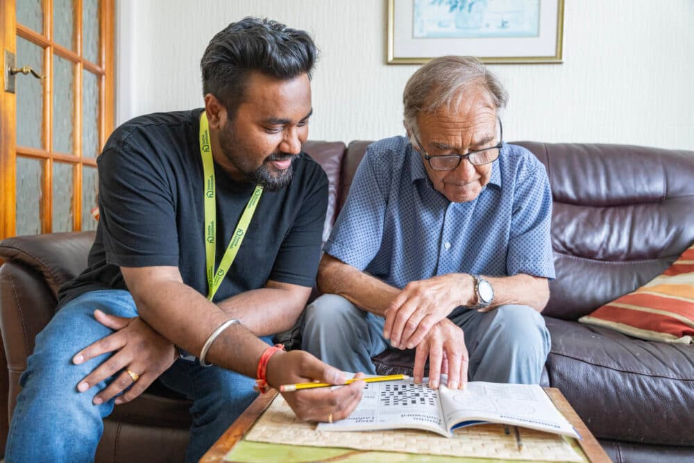 A younger man helps an older man solve a crossword puzzle on a coffee table in a living room. - Home Instead