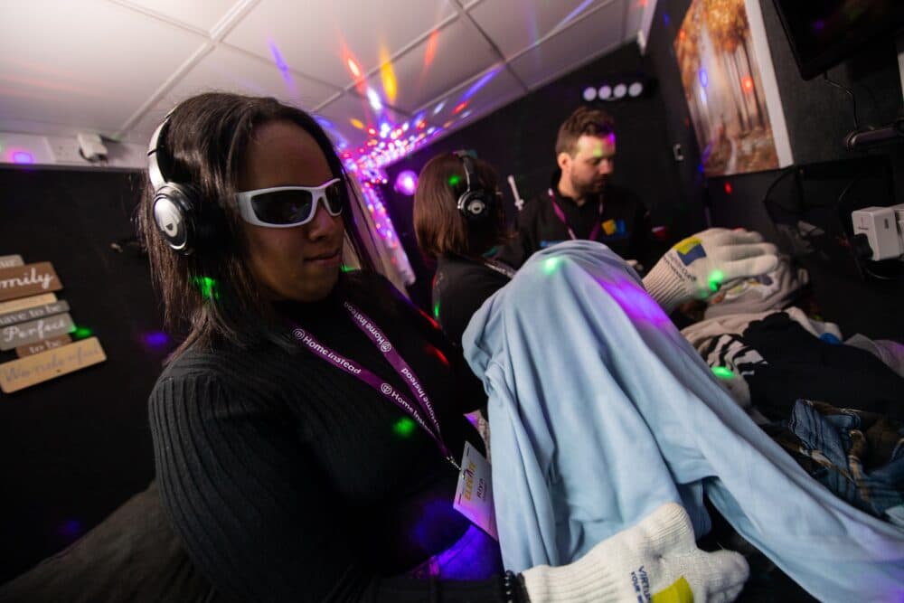 A woman wearing dark glasses, headphones, and gloves labeled "Virtual Dementia Tour" participates in a sensory simulation experience. She is holding a light blue shirt and appears focused. Two other individuals are in the background, also wearing headphones. The room is dimly lit with colourful light projections and has a cozy, home-like decor. One person is wearing a lanyard with the branding "Home Instead."