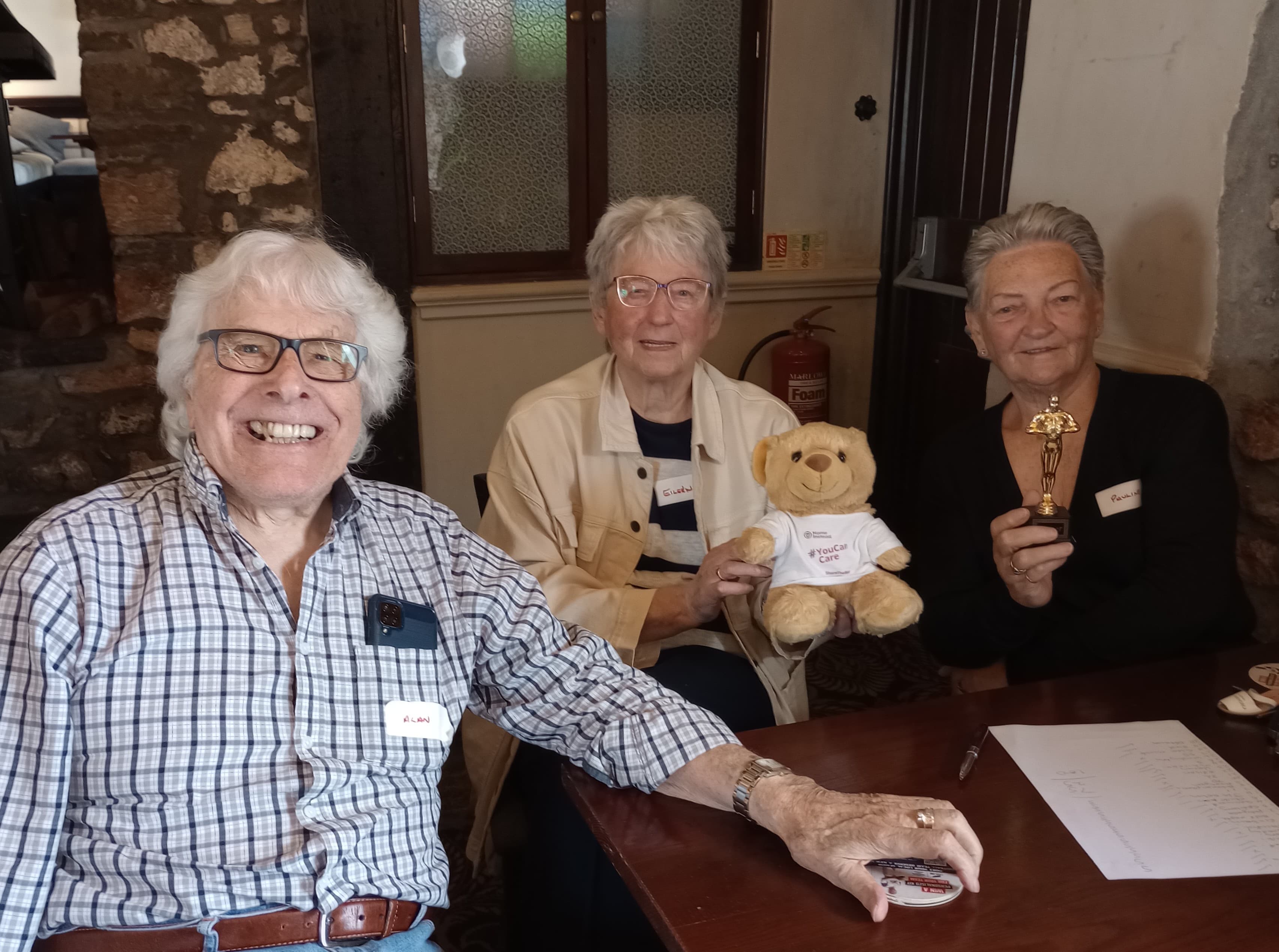 Three smiling elderly people sit at a table; one holds a teddy bear, another holds a small trophy. - Home Instead