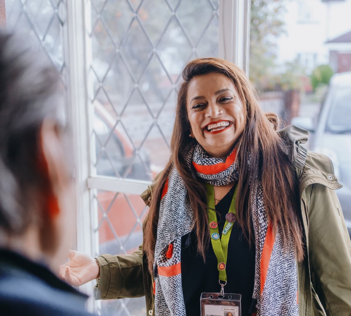A female carer with long straight hair wearing a scarf entering the door and happy and smiling