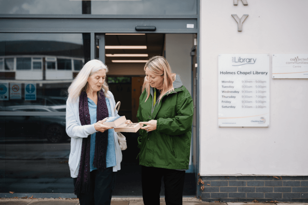 Two women stand outside a library, looking at books together and smiling near the entrance. - Home Instead
