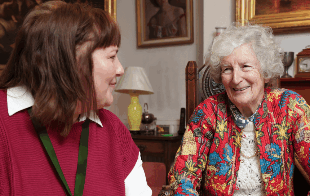 An elderly woman smiles while talking with a younger woman in a cozy, warmly decorated room. - Home Instead