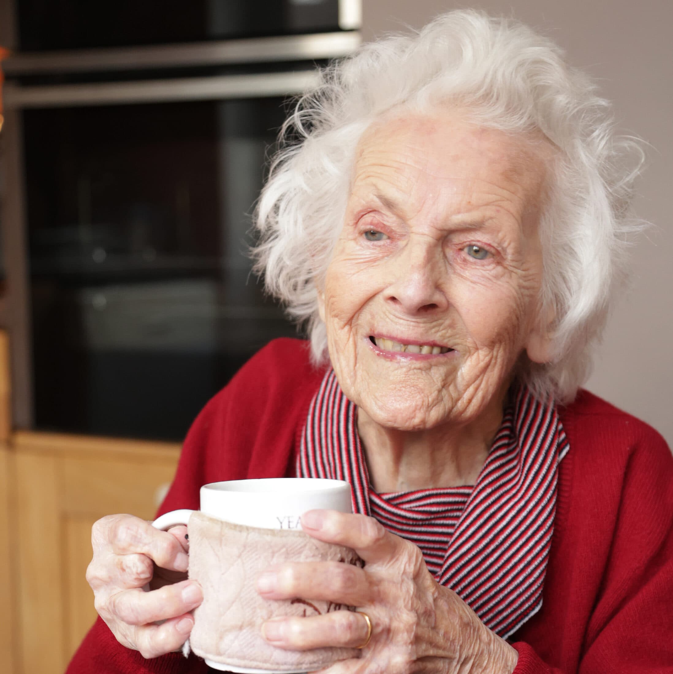 Smiling elderly woman in a red sweater holding a mug, sitting indoors. - Home Instead
