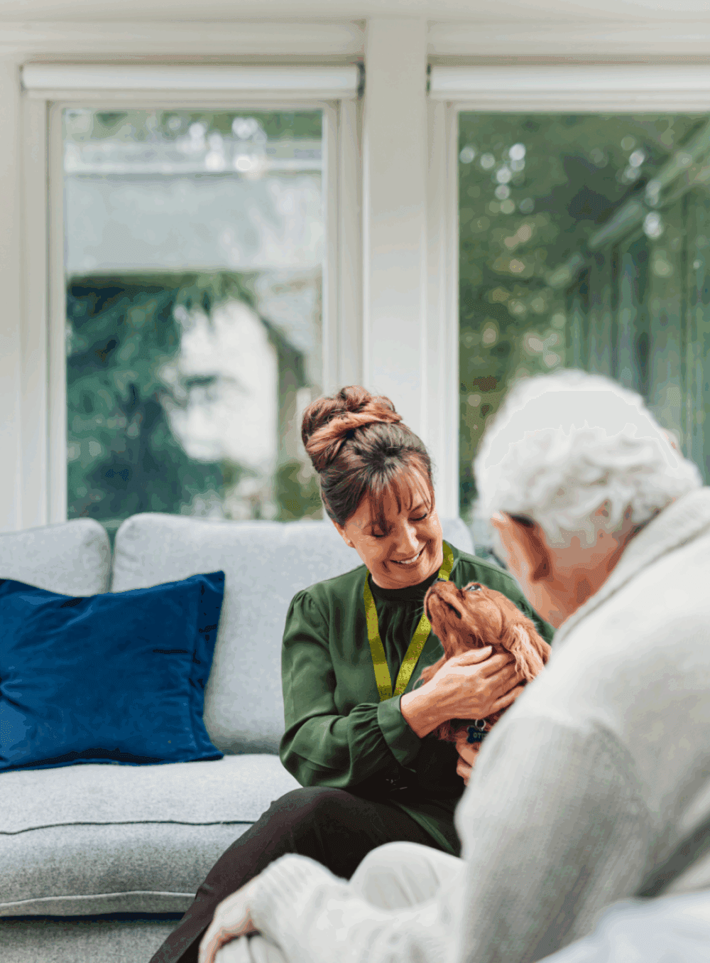 A woman smiles while holding a small dog, sitting next to an older man on a sofa in a bright room. - Home Instead