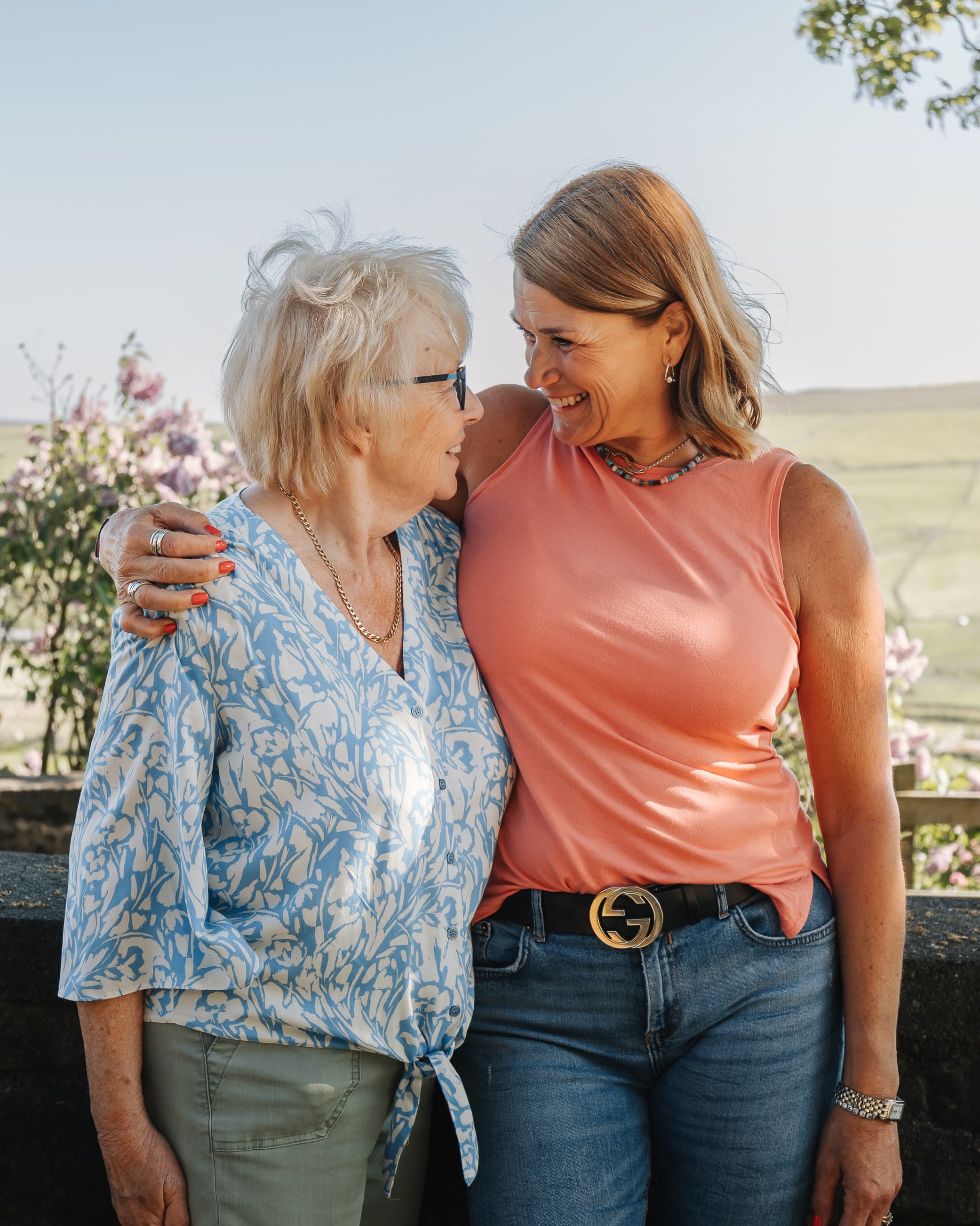 Two women smiling and embracing outdoors, standing closely and looking at each other warmly. - Home Instead