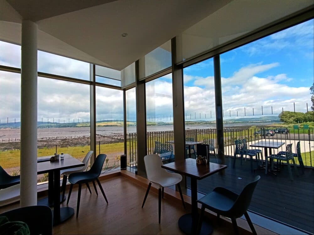Modern cafe with glass walls overlooks a scenic outdoor deck and expansive landscape with water and distant hills. - Home Instead