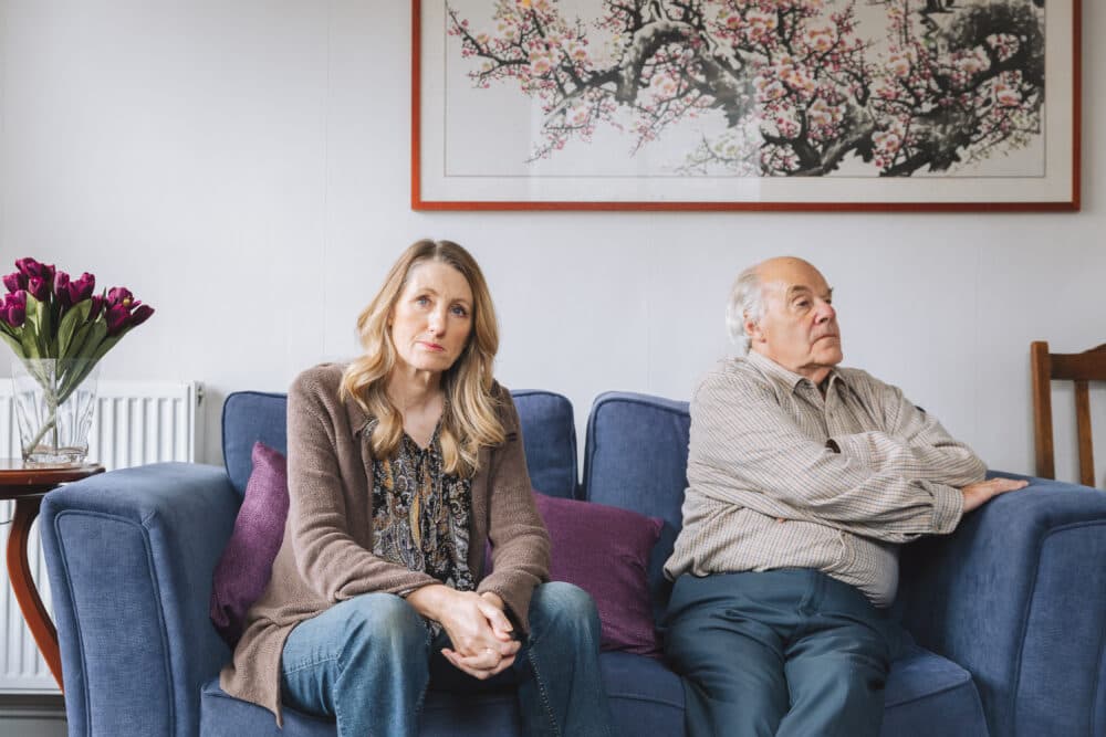 A woman and an older man sit on a couch, looking serious and facing away from each other. - Home Instead