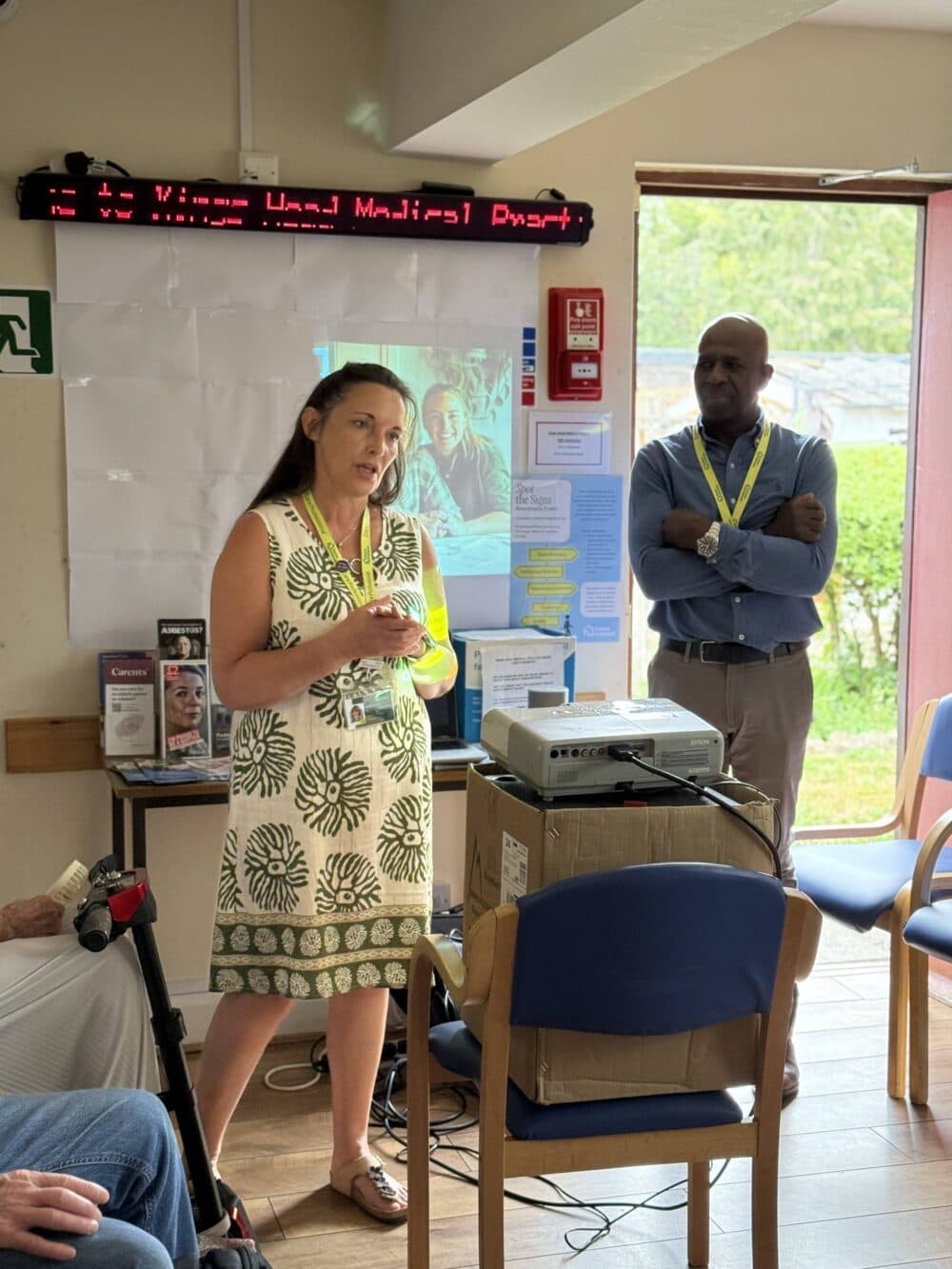 A woman presents to a small group indoors while a man stands nearby, listening attentively. - Home Instead