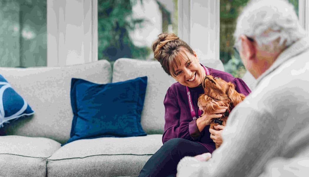 Smiling woman with a dog sits on a sofa, facing an elderly man in a bright living room. - Home Instead