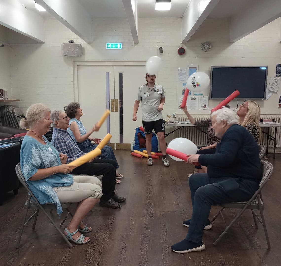 Six older adults sit in a circle hitting balloons with foam noodles, while an instructor stands nearby. - Home Instead