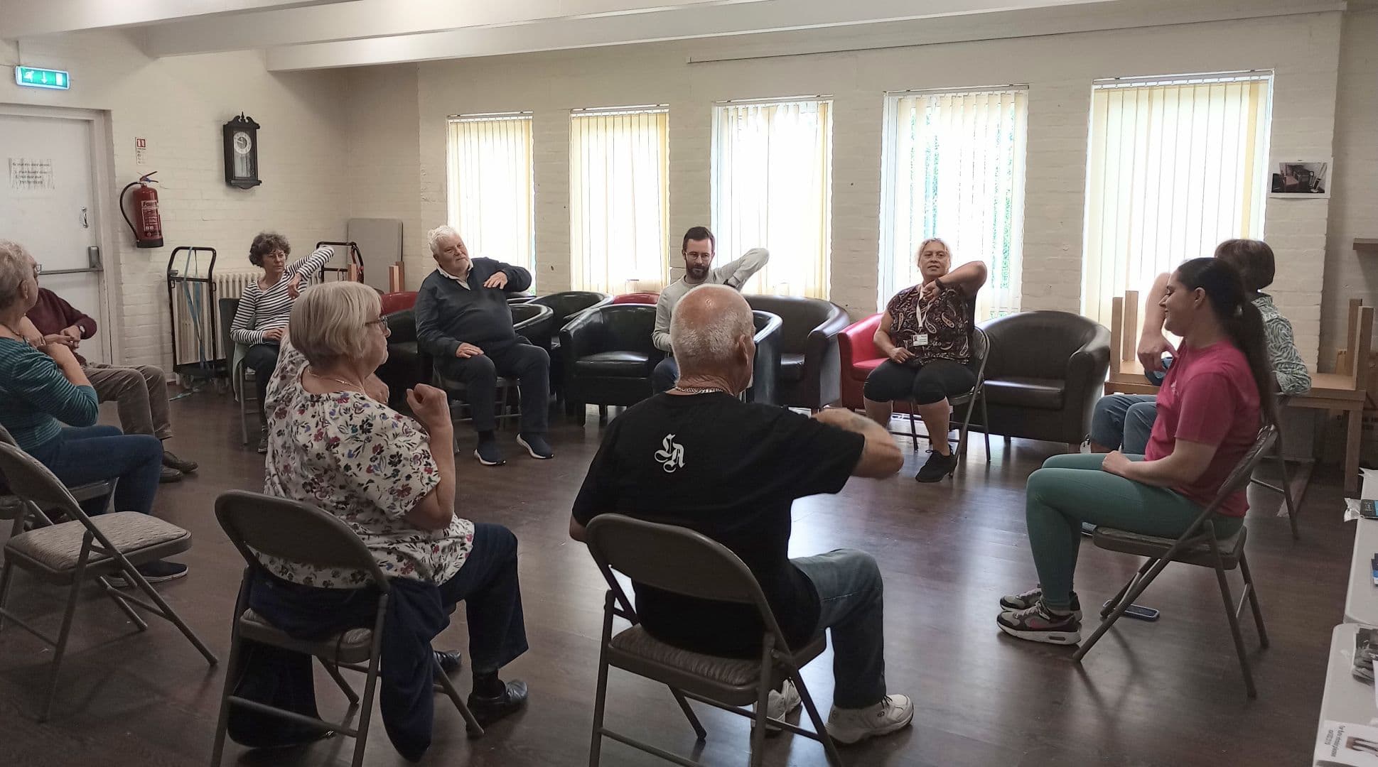 A group of people sit in a circle on chairs, participating in a seated exercise class indoors. - Home Instead