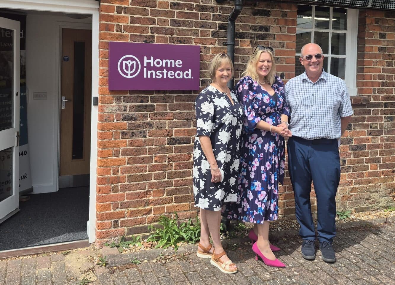 Three people are standing outside a brick building with a sign that reads "Home Instead." Two women and one man are smiling and posing for the photo; one woman is shaking hands with the man. The women are wearing floral dresses, and the man is in a checkered shirt with dark trousers. The building has white-framed windows and an open door with part of a banner visible inside. The setting appears to be sunny and welcoming.