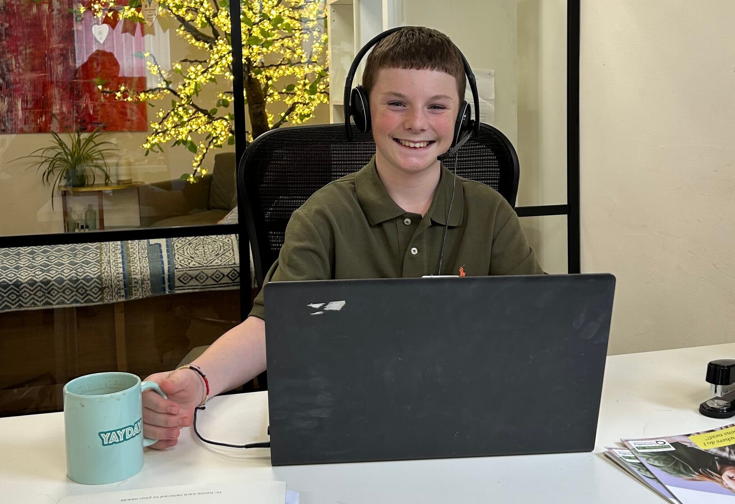 Smiling boy wearing headphones sits at a desk with a laptop and a "YAYDAY" mug in an office. - Home Instead