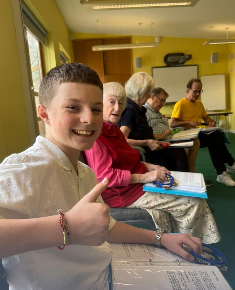 Teen boy gives thumbs up while sitting with older adults in a classroom holding papers and clipboards. - Home Instead