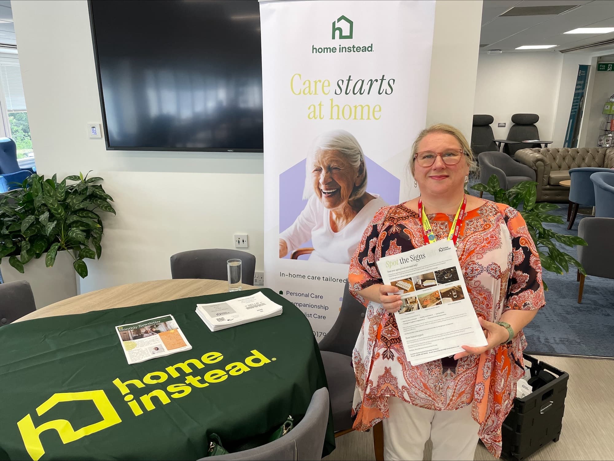Woman holding a leaflet at a "Home Instead" care booth with a promotional banner and table display. - Home Instead