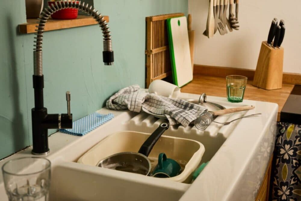A kitchen sink filled with dirty dishes and utensils, with a dish towel draped over the edge. - Home Instead