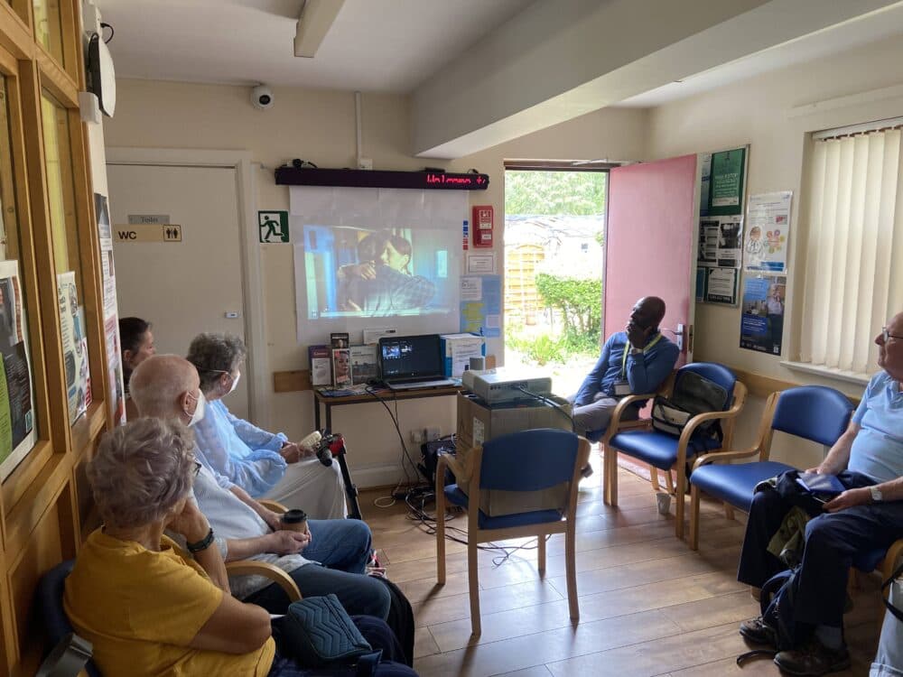 Several people sit in a waiting room watching a video projected onto a wall during the daytime. - Home Instead