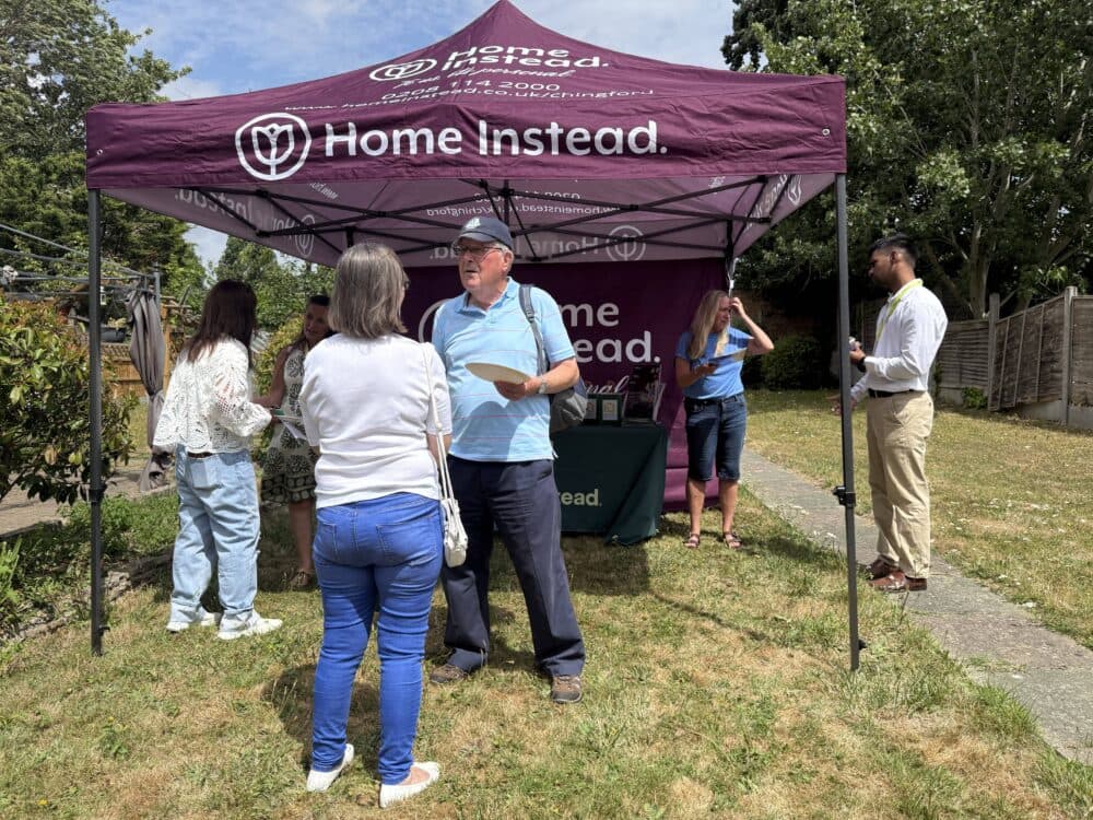 People gather and talk under a purple "Home Instead" tent at an outdoor event on a sunny day. - Home Instead