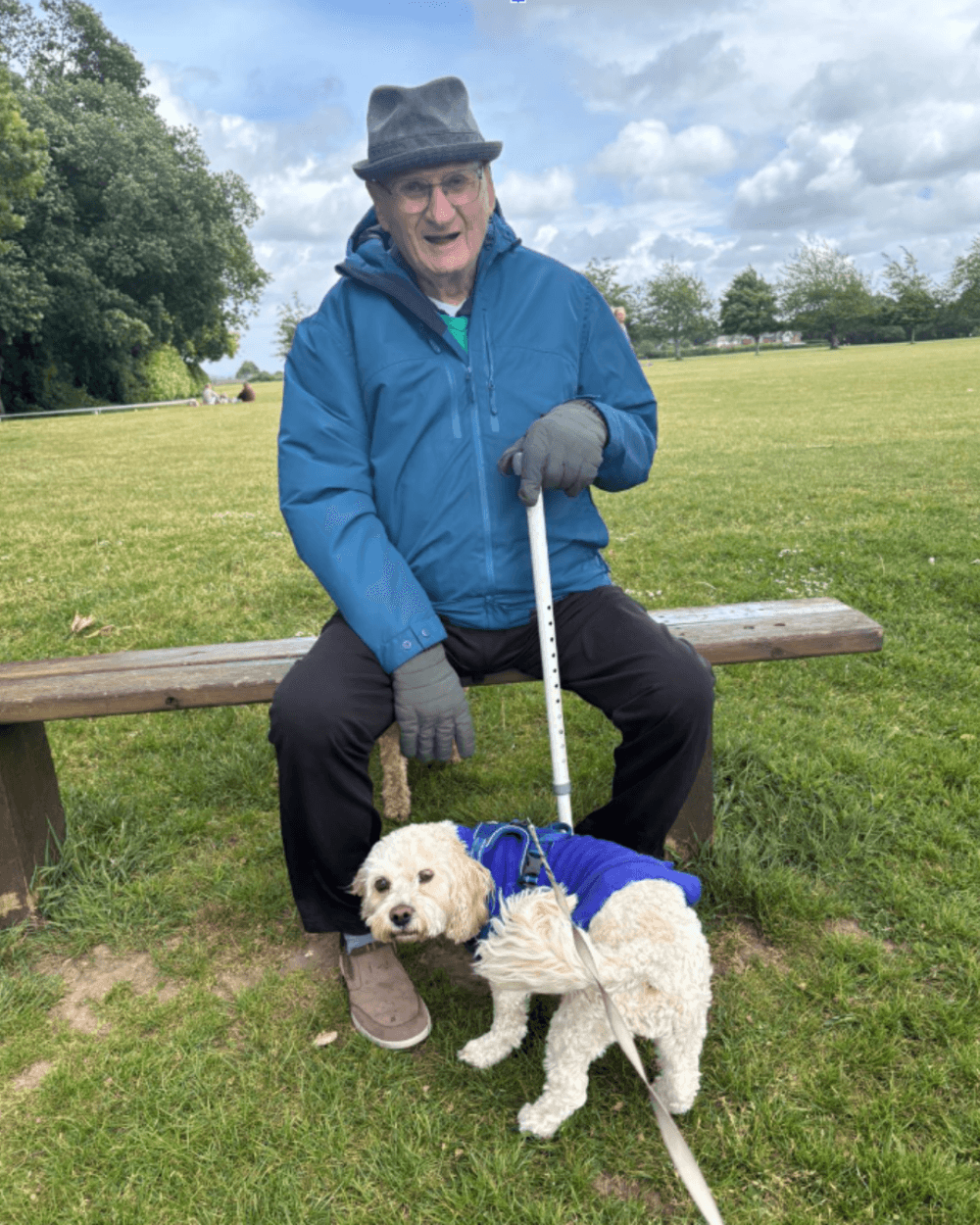 An older man in a blue jacket sits on a bench, holding a cane, with a small dog in a blue coat by his feet. - Home Instead