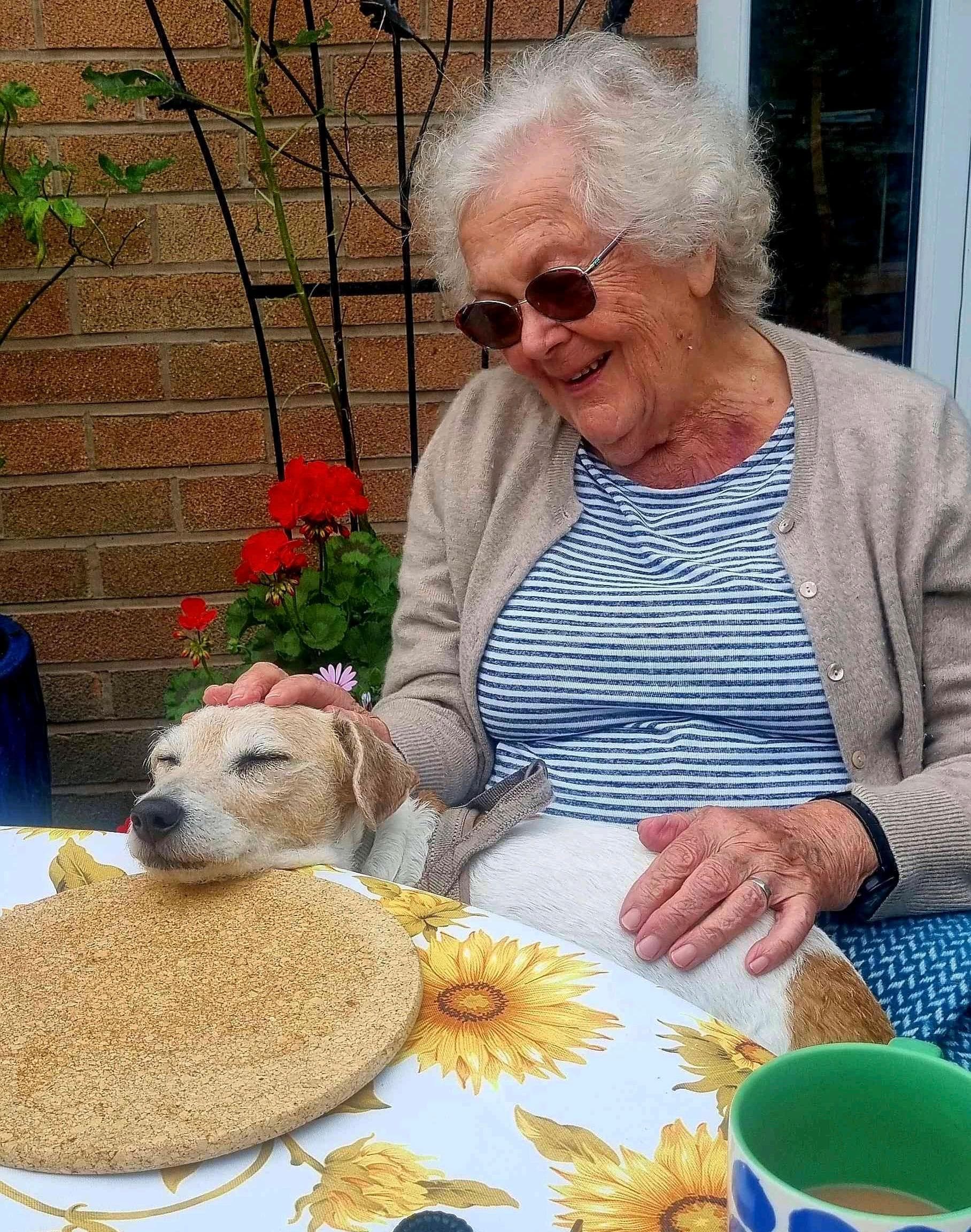 Smiling elderly woman pets a relaxed dog resting its head on a table with a sunflower tablecloth. - Home Instead