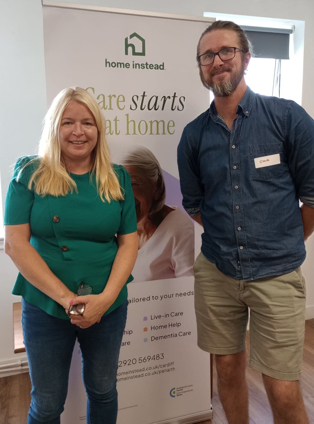 Two people smiling in front of a "Home Instead" care banner inside a brightly lit room. - Home Instead