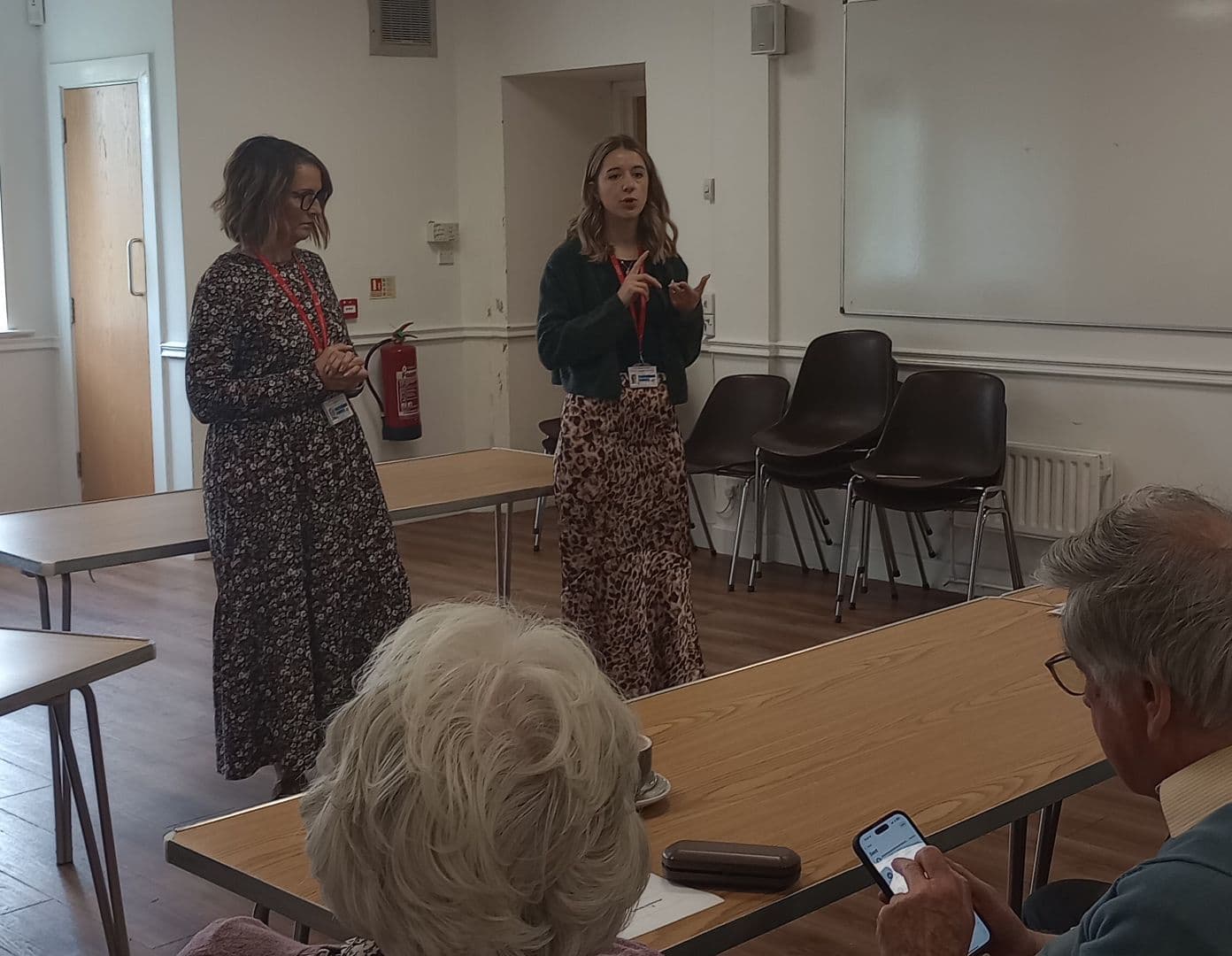 Two women stand and speak to older adults seated in a classroom with tables and chairs. - Home Instead