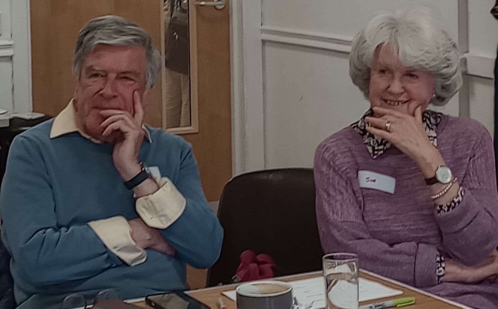 An older man and woman sit at a table, both smiling with their hands on their chins. - Home Instead