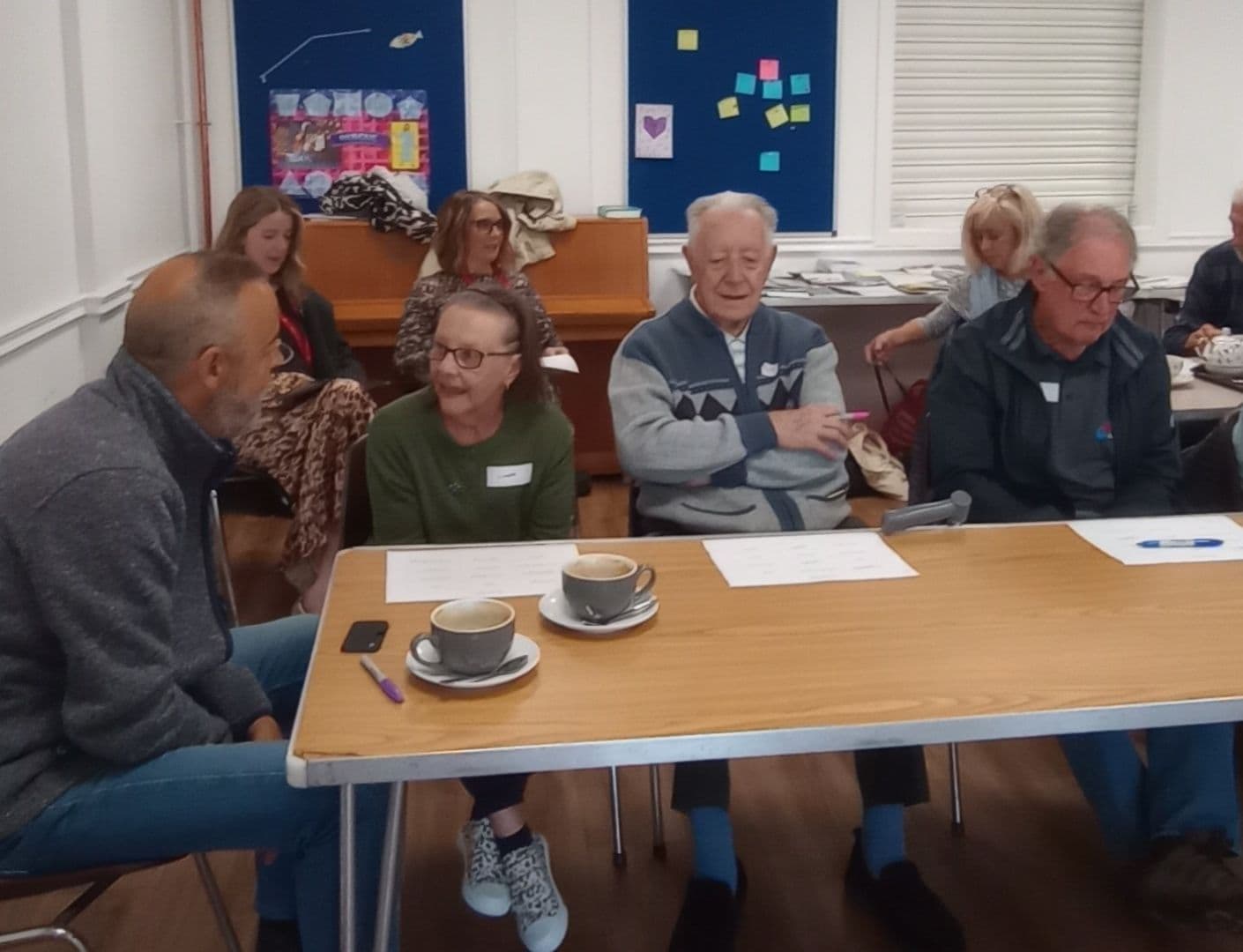 Four older adults sit at a table with cups, talking, in a community room with others in the background. - Home Instead