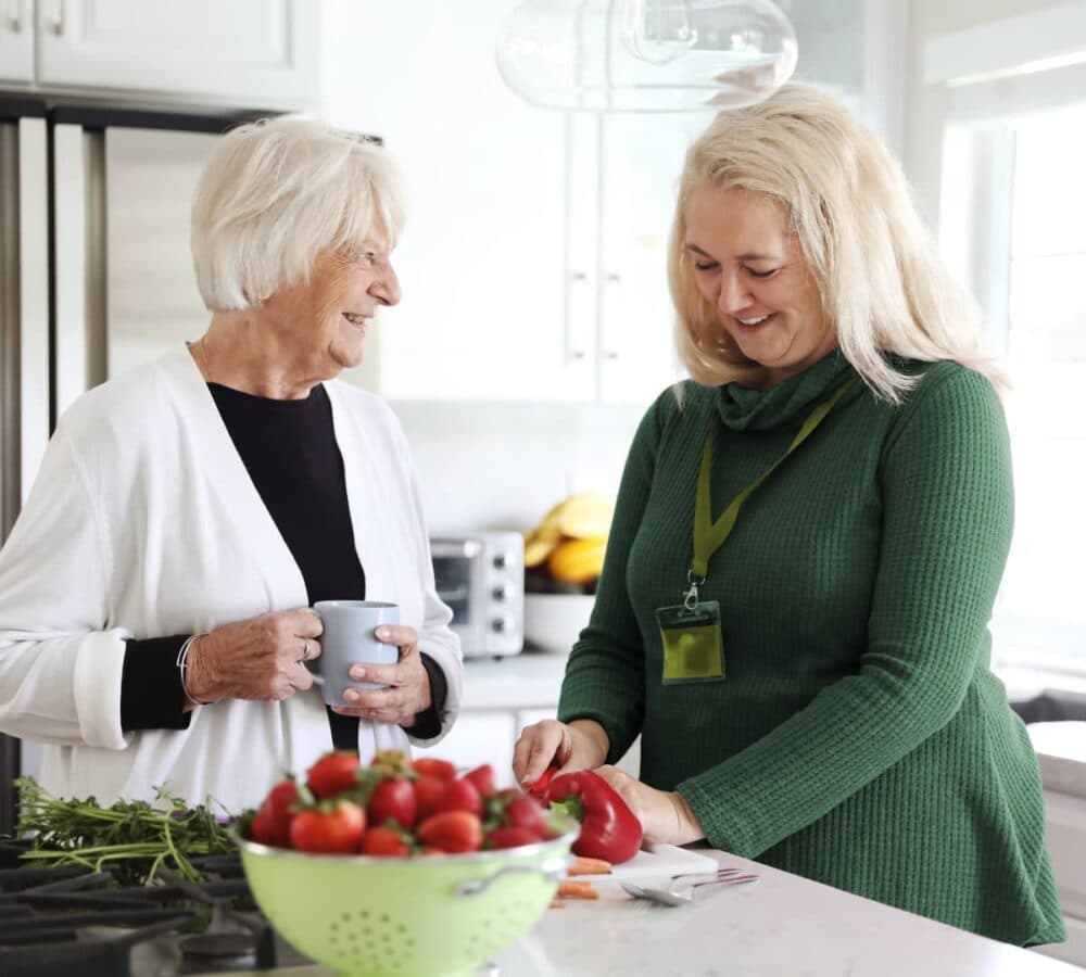 An older female adult having coffee with her younger female carer cutting vegetables