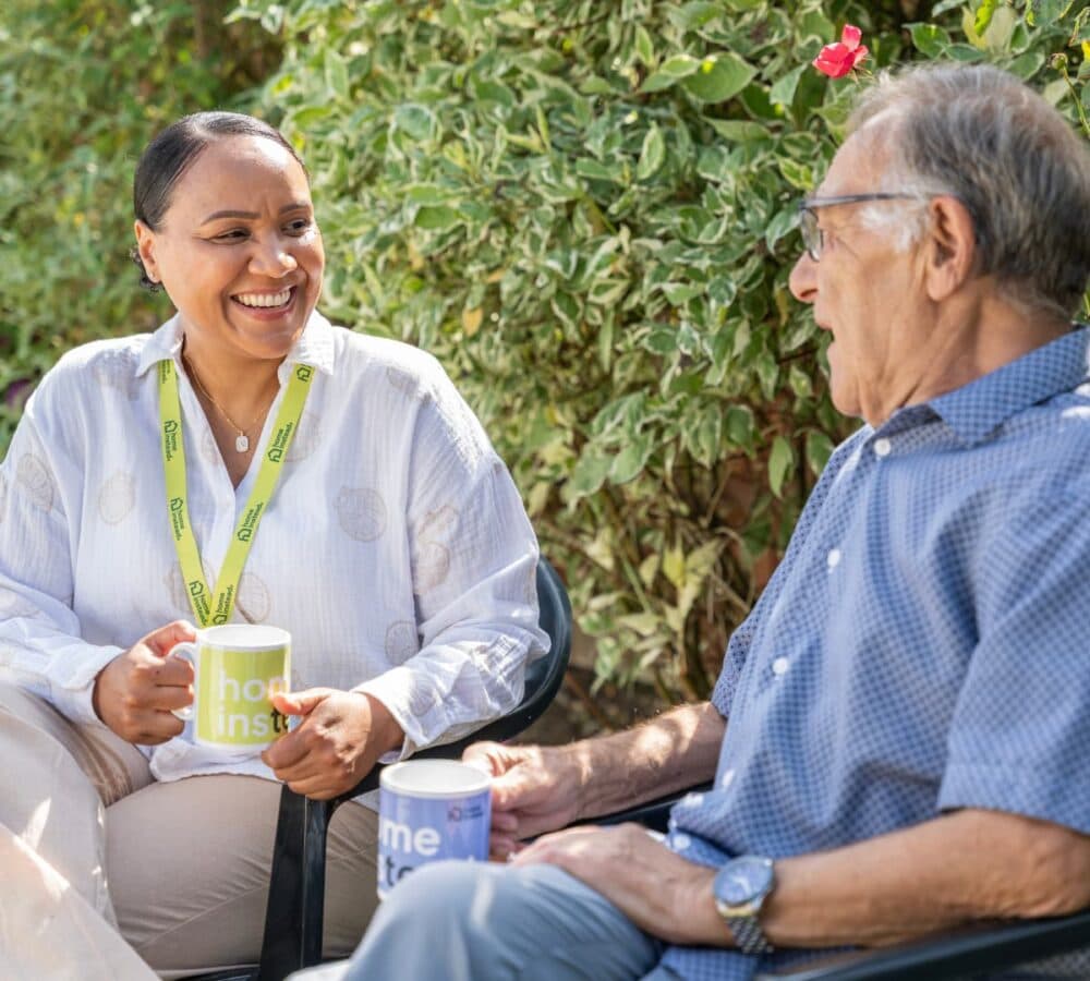 An older male adult with grey hair and wearing eyeglasses with a younger female carer both chatting and smiling while drinking coffee in the garden