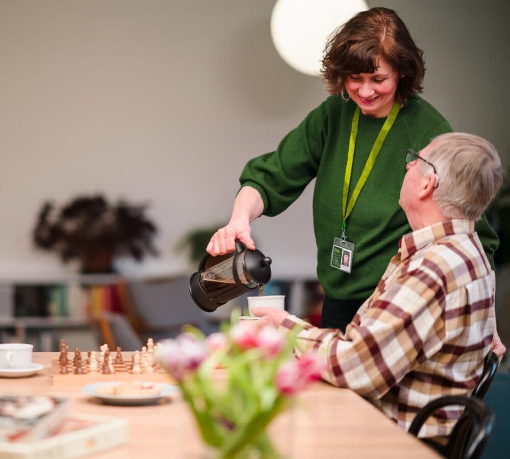 An older male adult with grey hair and wearing eyeglasses and stripe brown long sleeves sitting on a chair while having coffee and chatting with his female carer wearing green