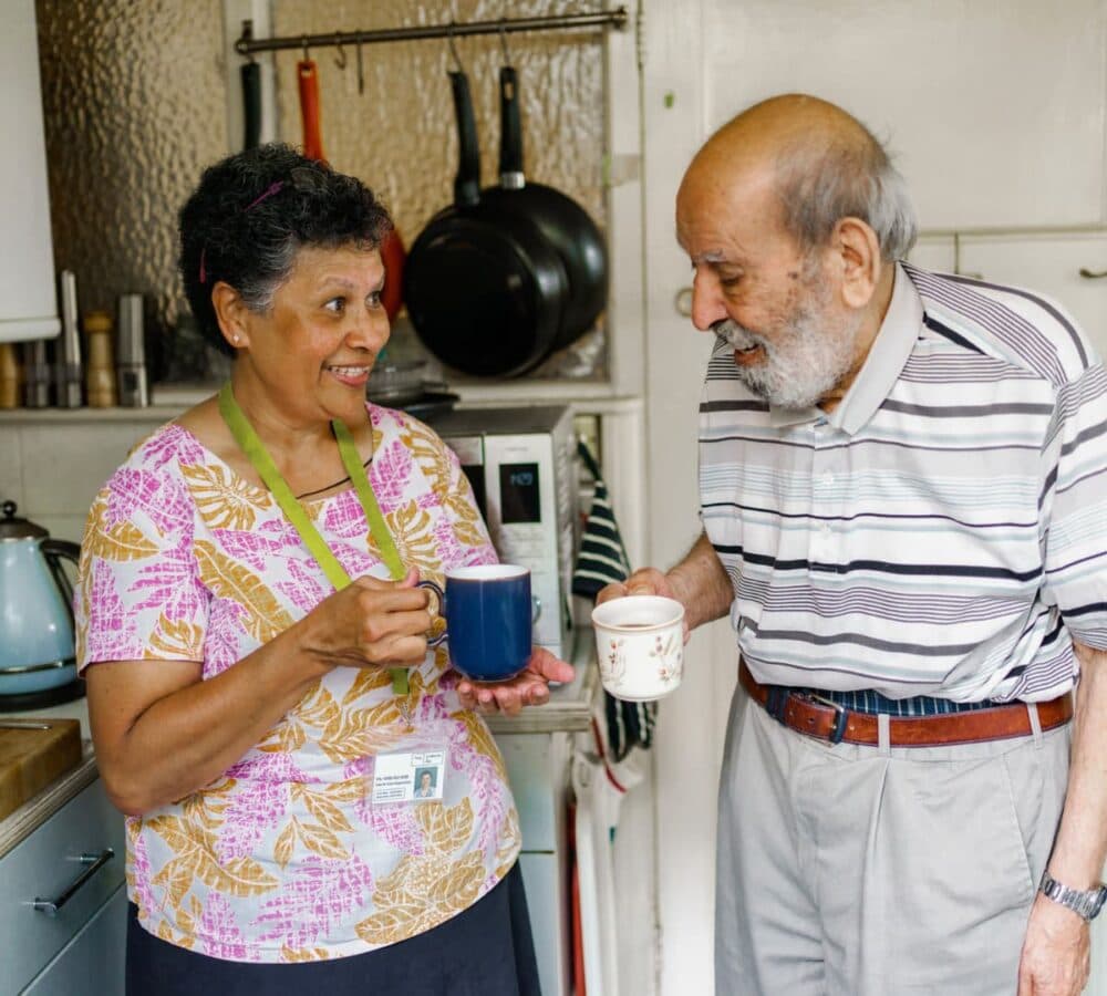 An older male adult with grey hair having a cup of coffee with his younger female carer inside the kitchen