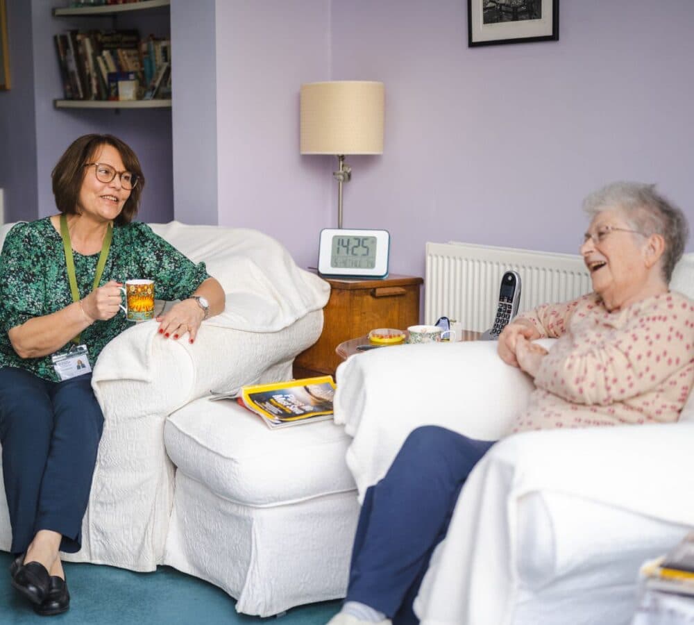 An older female adult with grey hait sitting on a white couch chatting with her youger female carer with short hair and wearing eyeglasses while drinking coffee