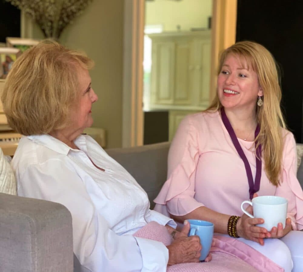 An older female adult with white long sleeve sitting on the couch while drinking coffee and having a chat with her younger female carer with long blonde hair and wearing pink inside the house