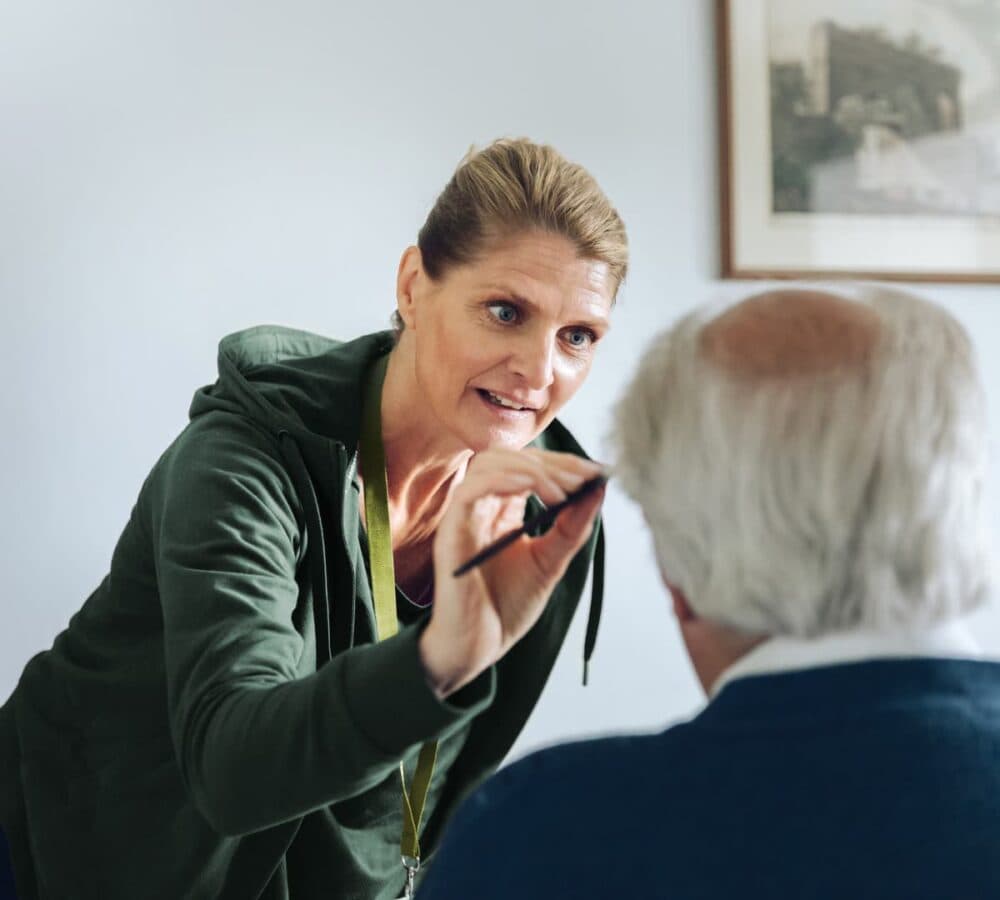 A young female carer helping an older male adult with grey hair and wearing blue inside his room