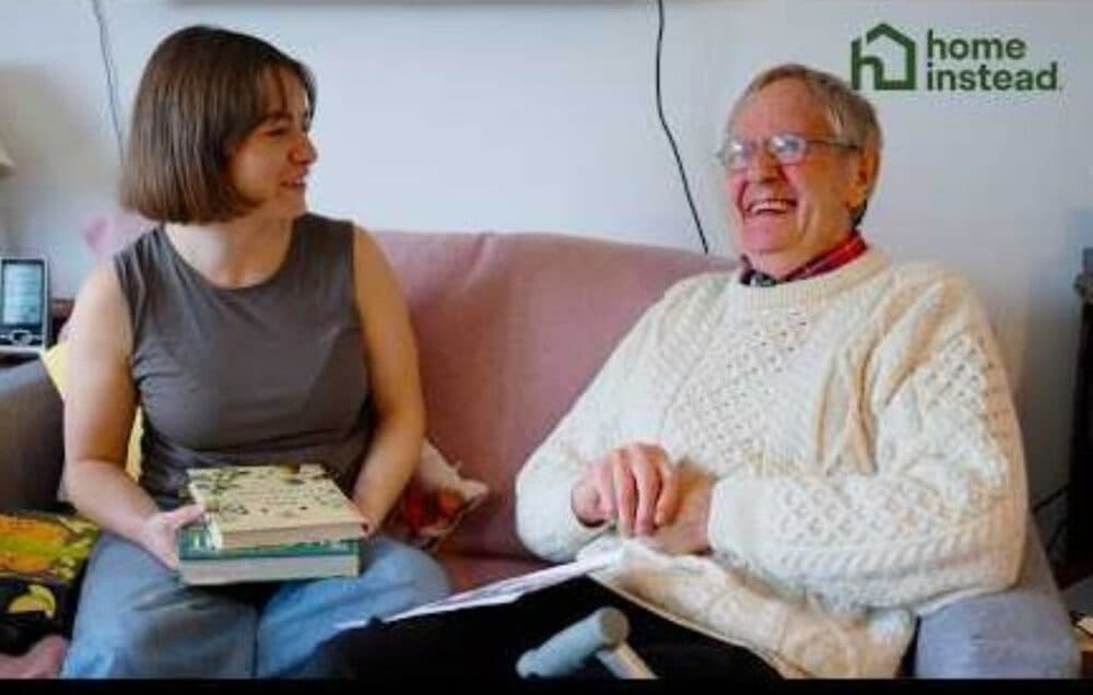 A young woman and an elderly man smile together on a sofa; she holds books, he uses a cane. - Home Instead