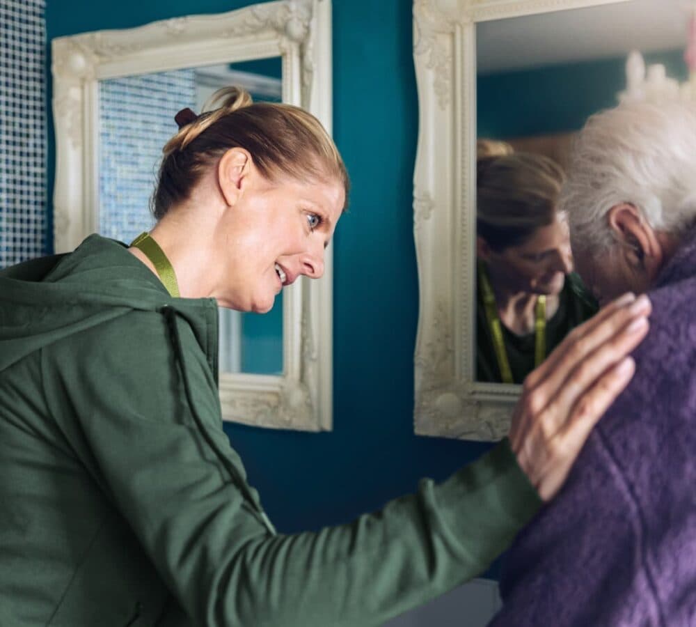 Caregiver comforting an elderly person, gently touching their shoulder in a home setting with mirrors. - Home Instead
