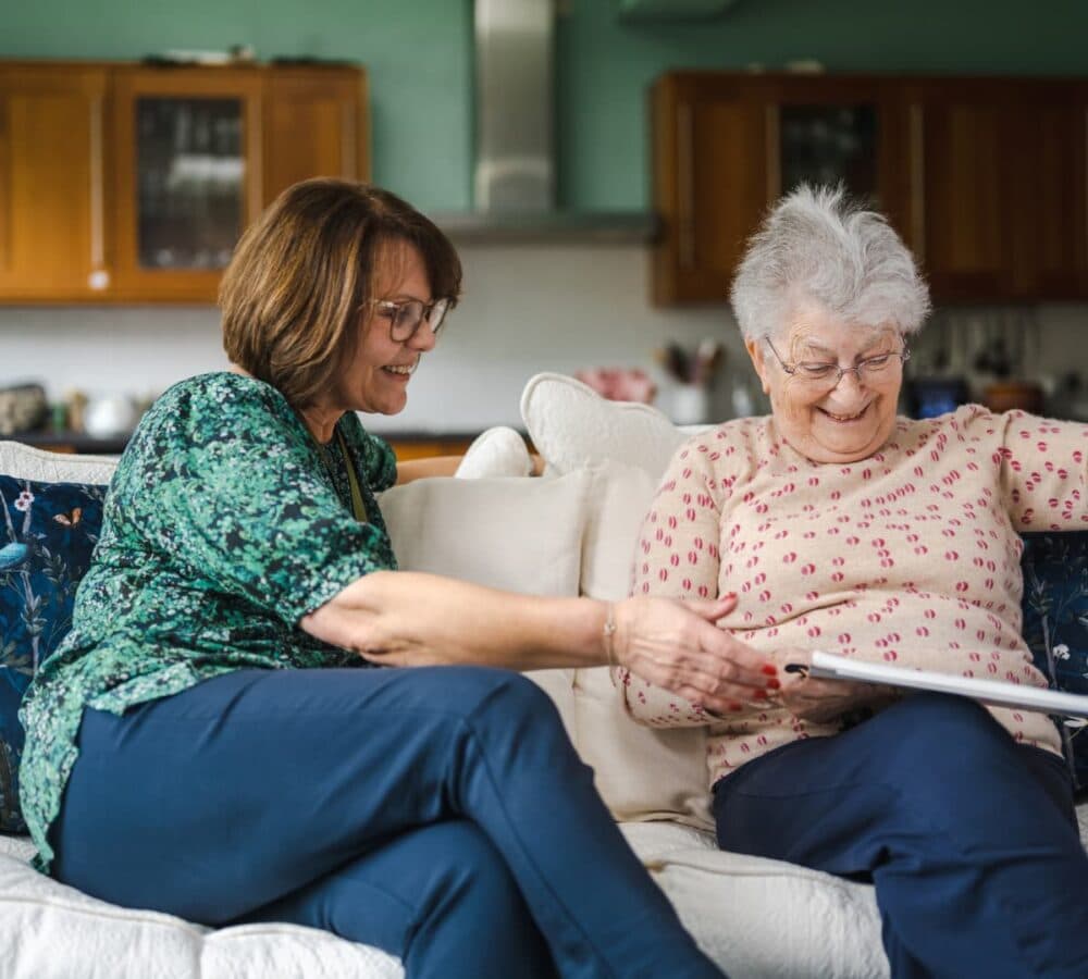 Two women looking at the album while happy and smiling and sitting on the couch inside the house