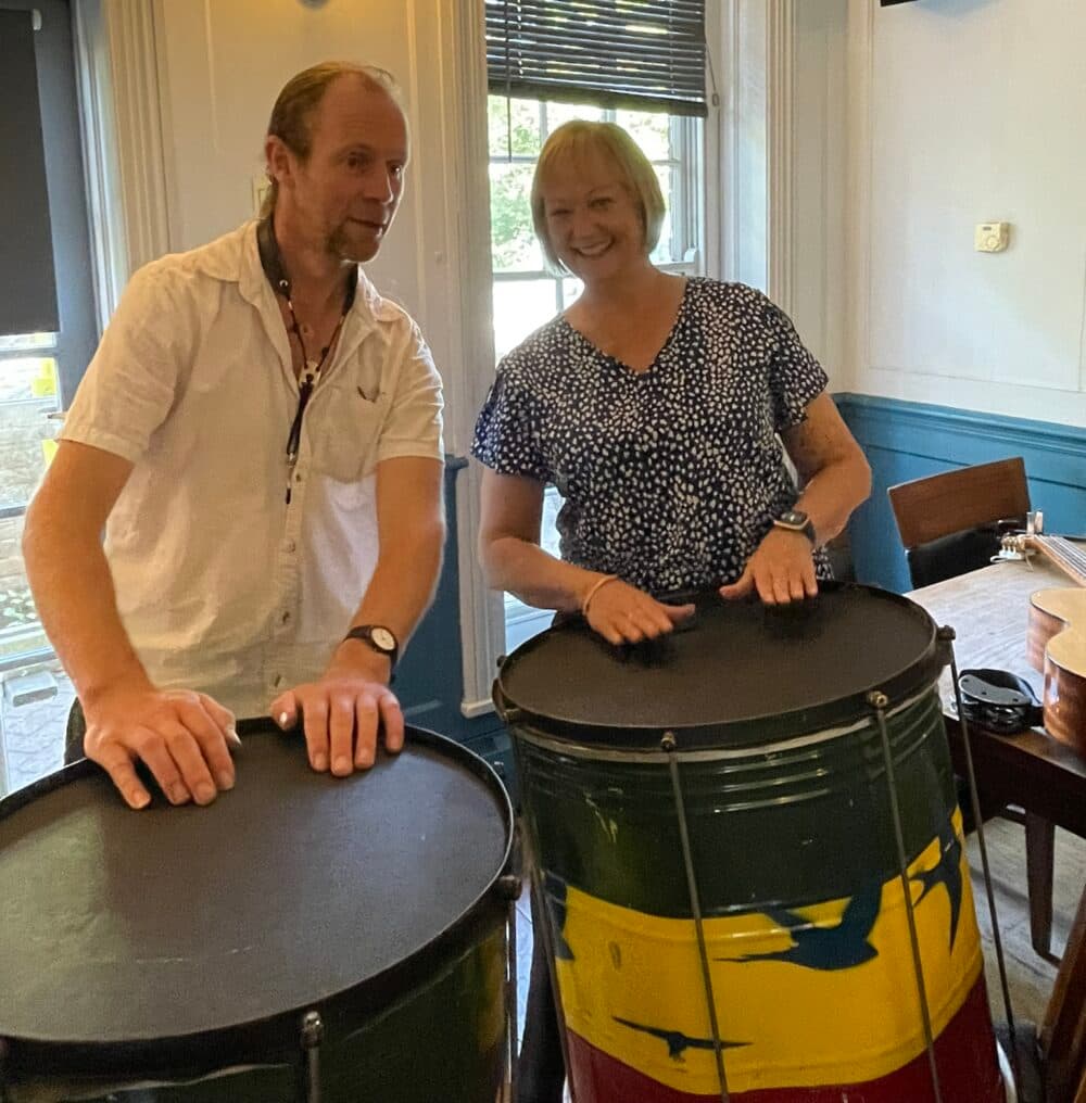 A man in a white shirt and woman in a black polka dot top stand smiling behind large colorful drums in an elegant room with white paneled walls, wall sconces, and venetian blinds. The drums appear to be steel drums or similar percussion instruments with green and yellow decorative patterns.