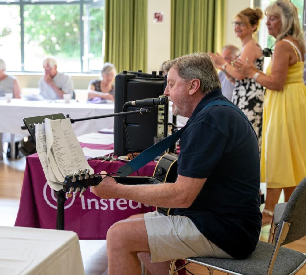 A man sitting while singing and playing the guitar inside the hall with notes ad lyrics in front of him
