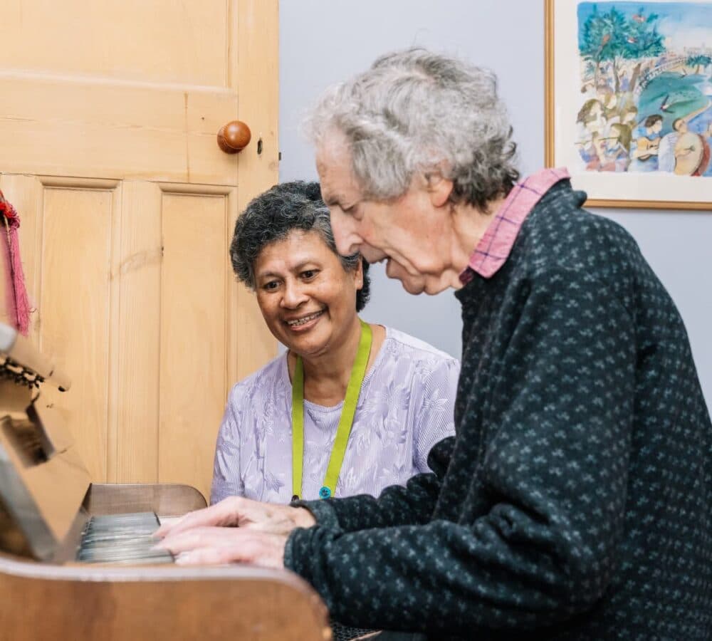 An older male adult with grey hair and playing the piano with his female carer with black hair happy and smiling