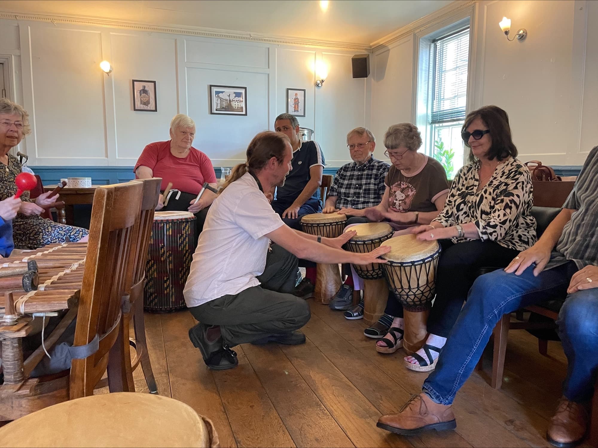 A group of older adults participating in a drum circle activity in what appears to be a community center or cafe. A man in a white shirt kneels in the center, facilitating the session while several participants sit in chairs holding various drums and percussion instruments. The room has blue and white painted walls with framed artwork, wall sconces, and large windows. On the left side, there's a wooden xylophone or marimba. The participants appear engaged and are learning to play their instruments together in this therapeutic or recreational music session.